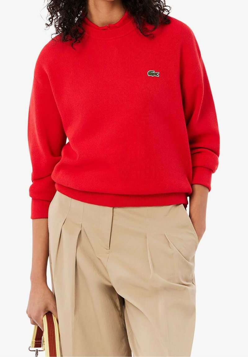 Personne portant un pull rouge Lacoste avec un pantalon plissé beige, tenant dans une main une bandoulière de sac à main beige et marron rayée.