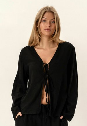 Blondínka v ľahko voľnom čiernom top s dlhým rukávom zviazanom vpredu, stojaca s rukami v vreckách na jednoduchom svetlom pozadí.