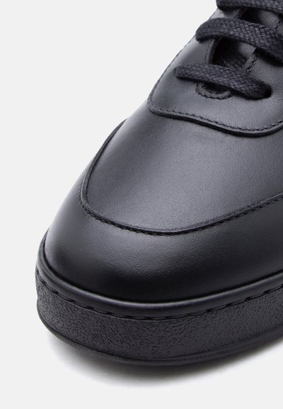 Κοντινή λήψη μαύρου δερμάτινου παπουτσιού με παχιά υφασμένη σόλα και μαύρα κορδόνια σε λευκό φόντο.