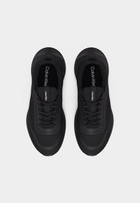 Ζευγάρι μαύρα αθλητικά παπούτσια Calvin Klein με κορδόνια, από πάνω σε λευκό φόντο.