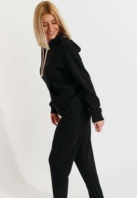 Schwarzer Kapuzenpullover mit pinken Kordeln, ausgestattet mit einer Kängurutasche und elastischen Bündchen, kombiniert mit passenden schwarzen Jogginghosen.