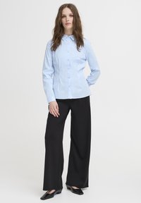 Svijetloplava košulja na dugme s ovratnikom, tijesnim strukom i dugim rukavima, u kombinaciji s crnim širokim hlačama visokog struka i crnim cipelama.