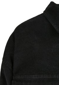 Tissu en velours côtelé noir avec une texture côtelée verticale, présentant un col et une coupe ajustée. Vue rapprochée de la zone de l'épaule du vêtement.