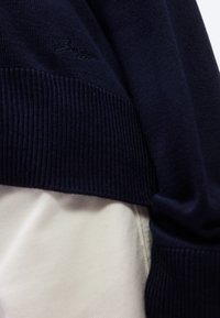 Κοντινό πλάνο στο μανίκι και τη μέση ενός πουλόβε ρ πλεκτού σε ναυτικό μπλε χρώμα πάνω σε ύφασμα ανοιχτό μπεζ παντελονιού.