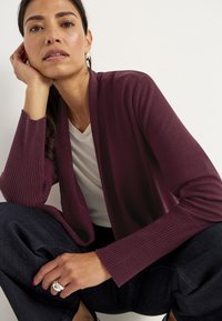 Vrouw met donker haar, die zilveren oorringen, een kastanjebruine cardigan, een wit shirt en donkere broek draagt, haar hoofd op haar hand steunt.