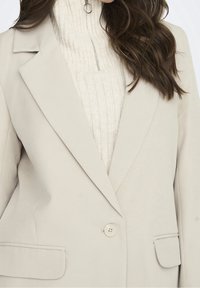Blazer beige claro con cierre de un solo botón, con solapas de muesca y dos bolsillos frontales. Llevado sobre un suéter de cuello alto de canalé crema.