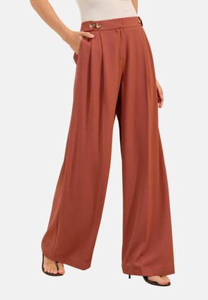 Donna che indossa pantaloni a gamba larga a vita alta color ruggine con dettagli a bottoni, abbinati a tacchi neri open-toe e top bianco.