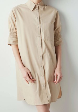 Persona que lleva un vestido camisa beige con botones, mangas cortas abullonadas con detalle fruncido y un bolsillo en el pecho, de pie contra una pared blanca.