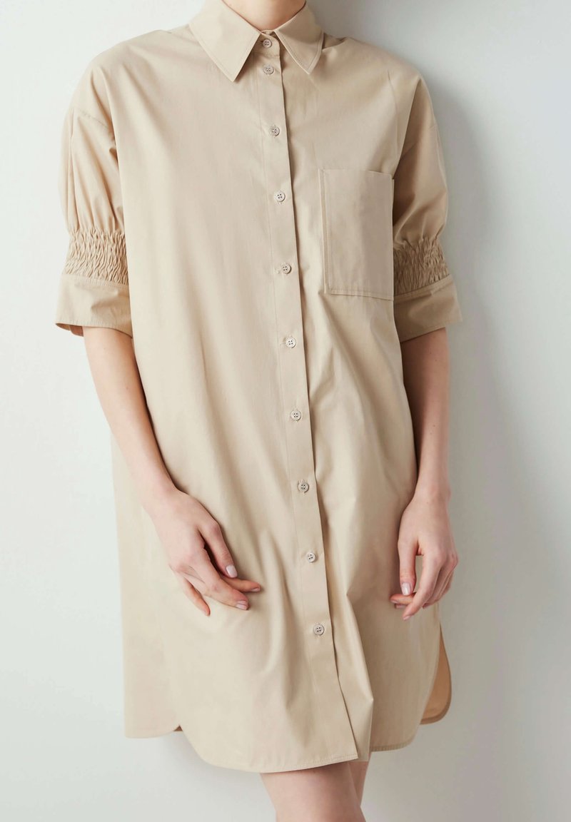 Personne portant une robe-chemise beige à boutons avec de courtes manches bouffantes présentant un détail smocké et une poche poitrine, debout contre un mur blanc.