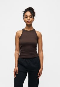 Jeune femme aux cheveux bouclés attachés, portant un haut marron sans manches et un pantalon noir, debout devant un fond blanc uni.