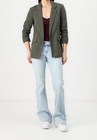 Blazer ajusté vert olive avec des revers, deux poches avant, associé à un jean évasé bleu clair et des baskets blanches.