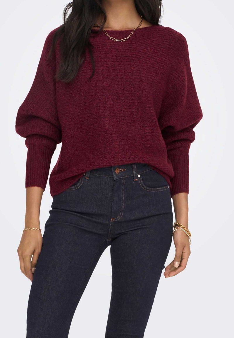 Burgundský pletený svetr s oversize rukávy, zaobleným výstřihem a strukturovaným materiálem. V kombinaci s tmavě modrými džínami s vysokým pasem.