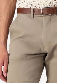 Beige bukser med en glat tekstur, der har en klassisk pasform, bæltestropper og et brunt læderbælte. Bemærkelsesværdig syning langs sømmen.