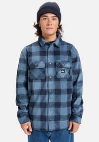 Camisa de franela a cuadros azules que presenta un cierre frontal de botones, dos bolsillos en el pecho con cierres a presión y una etiqueta de tela en el bolsillo izquierdo.
