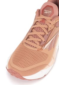 Bežecká obuv Reebok s svetloružovým sieťovaným zvrškom, texturovanými prekrývaniami a zodpovedajúcimi šnúrkami. Obsahuje bielu polstrovanú podrážku a akcenty loga.