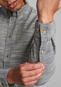Camicia grigia con bottoni, caratterizzata da un motivo a righe strutturato. Presenta un colletto arrotondato e polsini con bottoni di colore chiaro.
