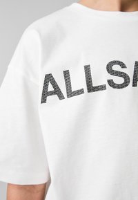 Vit bomulls t-shirt med fet, svart tryckt text som läser "ALLSAINTS" i ett upprepat mönster. Avslappnad passform, rundad krage, korta ärmar.