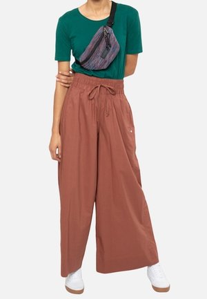 Pantaloni a vita alta dal taglio ampio di colore ruggine con tasche laterali, abbinati a una maglietta a maniche corte color teal e a una borsa a tracolla multicolore.