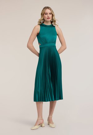 Nainen, jolla on päällään hihaton, vihreäsininen laskostettu midi-mekko sivuaukkoineen ja beige jykevä korkokengät, seisoo kädet selän takana.