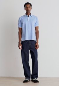Camisa polo azul clara com colarinho branco, combinada com calças de ganga escura de corte relaxado e ténis pretos com detalhes brancos.
