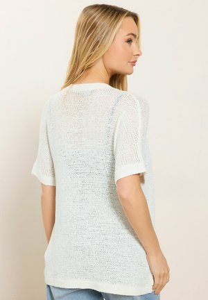 Blonde Frau, die ein locker sitzendes weißes Strickoberteil mit kurzen Ärmeln trägt, seitlich stehend vor einem schlichten hellen Hintergrund.