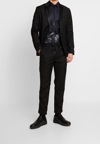 Giacca nera con rever in satin sopra una camicia a fantasia scura. Pantaloni neri a vestibilità rilassata con coulisse. Completato con sneakers nere.