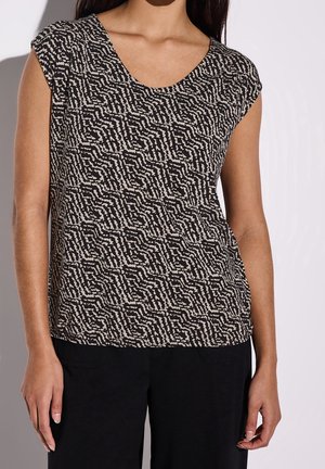 Kobieta w krótki rękaw, czarno-beżowej bluzce we wzory z okrągłym dekoltem, połączonej z czarnymi spodniami na jednolitym tle.
