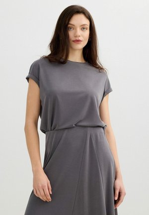 Mujer con cabello largo y oscuro que lleva un vestido gris suelto de manga corta, de pie frente a un fondo claro y sencillo.