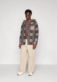 Καρό μπουφάν σε καφέ, ναυτικό μπλε και μπεζ με πράσινη κουκούλα και τσέπη στο στήθος, φορεμένο πάνω από καφέ πουκάμισο και μπεζ παντελόνι, σε συνδυασμό με λευκά αθλητικά παπούτσια.