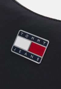 Logo di Tommy Jeans su uno sfondo in tessuto nero. Il logo presenta un design rettangolare con blocchi rossi e bianchi e include il nome del brand in blu navy.