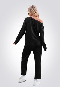 Felpa nera scivolata con maniche lunghe abbinata a pantaloni neri. Il modello indossa sneakers bianche, mostrando un fit rilassato e una texture morbida.