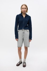 Suéter de punto acanalado navy con cremallera, pantalones cortos de mezclilla gris claro, zapatos marrones de patrón y calcetines blancos. Diseño sencillo, ajuste relajado y atuendo casual.