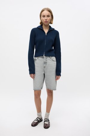 Pull en maille côtelée bleu marine à zip, short en denim gris clair, chaussures marron à motifs et chaussettes blanches. Design simple, coupe décontractée et tenue décontractée.