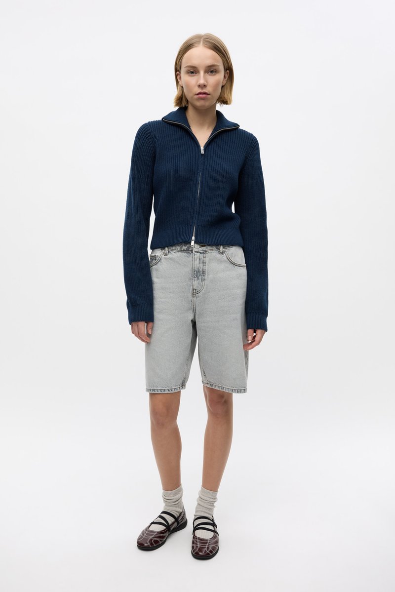 Pull en maille côtelée bleu marine à zip, short en denim gris clair, chaussures marron à motifs et chaussettes blanches. Design simple, coupe décontractée et tenue décontractée.