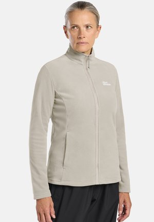 Vrouw draagt een lichtbeige fleecejack met rits en hoge kraag, staand tegen een effen witte achtergrond.