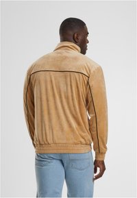 Bež žametna jakna z mimogrednim ovratnikom, elastičnimi manšetami in robom. Opremljena je z black obrobo ob ramenih in straneh.