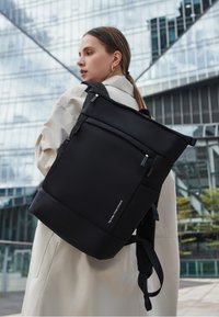 Czarny plecak o minimalistycznym designie, wykonany z gładkiego tworzywa sztucznego, z regulowanymi paskami i subtelnym logo marki.