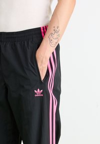 I pantaloni sportivi neri presentano un design a tre strisce rosa lungo i lati, una cintura elasticizzata e una piccola tasca con cerniera.
