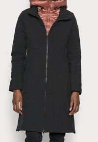 Abrigo negro con cierre frontal, que presenta un alto cuello y mangas de tres cuartos. Debajo, se puede ver una chaqueta acolchada de color rosa.