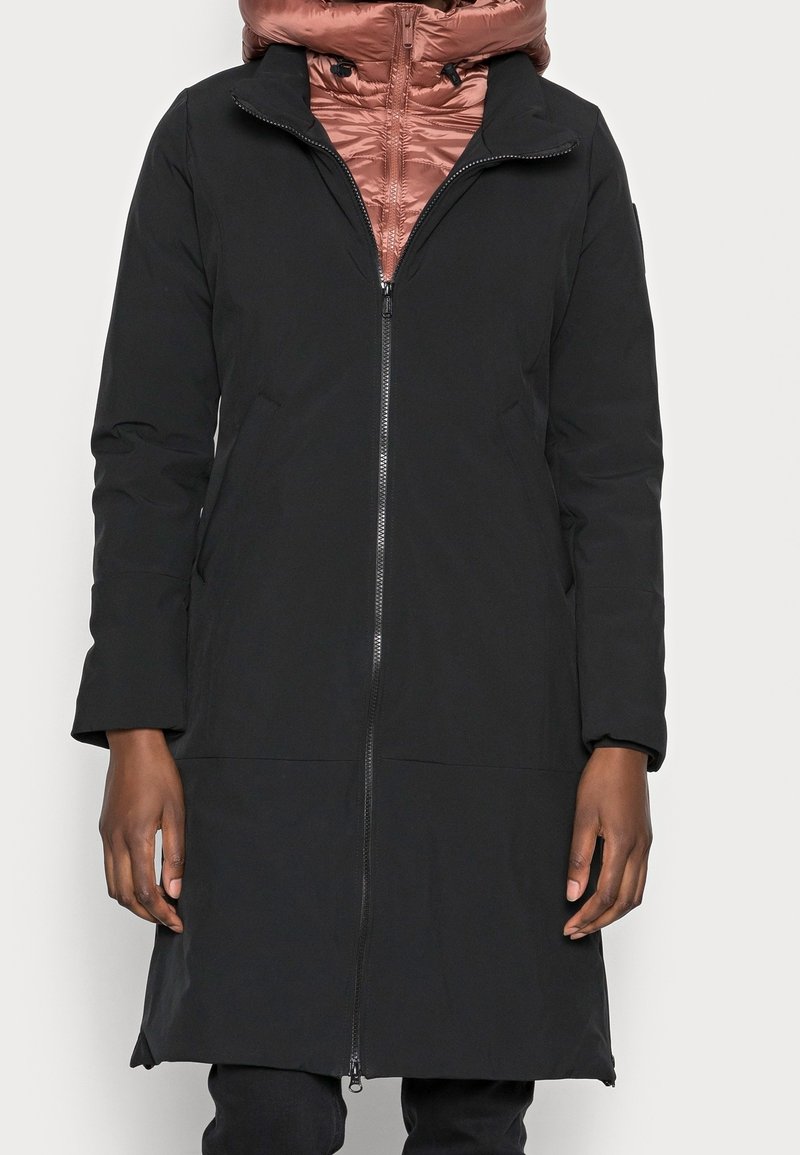 Abrigo negro con cierre frontal, que presenta un alto cuello y mangas de tres cuartos. Debajo, se puede ver una chaqueta acolchada de color rosa.