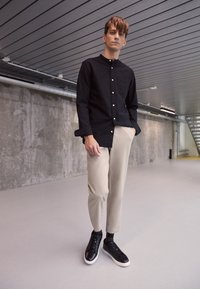 Black button-down shirt with a pocket paired with beige tapered pants and black sneakers with white soles, set against a concrete background.