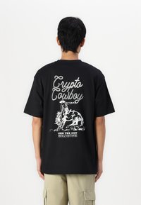 Sort bomulds t-shirt med et grafisk print på ryggen. Teksten siger "Crypto Cowboy" med en cowboy på en tyr i hvid. Løs pasform.