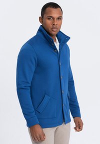 Sudadera azul con cuello alto, cierre de botones y dos bolsillos frontales. Textura suave y diseño ajustado.