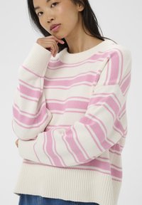 Jeune femme aux longs cheveux noirs portant un pull blanc à rayures horizontales roses, la main reposant près du menton, sur un fond uni.