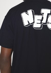 Camiseta de algodón negra con un gran gráfico blanco "NETS" en la parte trasera. Tiene cuello redondo y mangas cortas. Textura suave.