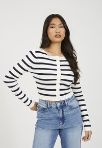 Biały kardigan w paski o kroju krótkim, z granatowymi paskami, z okrągłym dekoltem i zapięciem na guziki z przodu. Połączony z dżinsami z wysokim stanem.