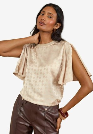 Kvinde iført beige kortærmet bluse med prikker og mørkebrune læderbukser med armbånd på højre håndled, poserer med venstre hånd bag nakken.