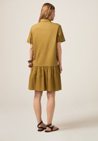 Rochie scurtă de culoare verde măsliniu, cu mâneci scurte, guler și fustă plisată. Textură netedă, până la genunchi, asortată cu sandale negre cu sireturi și brățări.