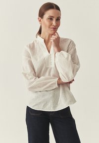 Femme portant un chemisier blanc à manches longues avec des motifs de feuilles et un jean foncé, debout, une main posée sur le menton et l'autre croisée.