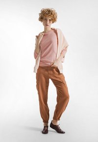 Jasnoróżowa koszula z długim rękawem, nałożona na jasnoróżowy sweter, w połączeniu z ceglasto-rudymi spodniami cargo ze ściągaczami oraz ciemnymi butami.